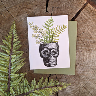 Greeting card | skull planter - Holt x Palm -  skull planter An archival print of various fern specimens, housed in a skull vase with tiki vibes. Reproduced from original illustrations by ilikesara / Sara Schalliol-Hodge. -Card measures 4.25" x 5.5" when folded -Printed on 100 lb off-white cover paper -Avocado green envelope included -Blank interior -Packaged in a clear cellophane sleeve Designed and printed in Denver, Colorado.